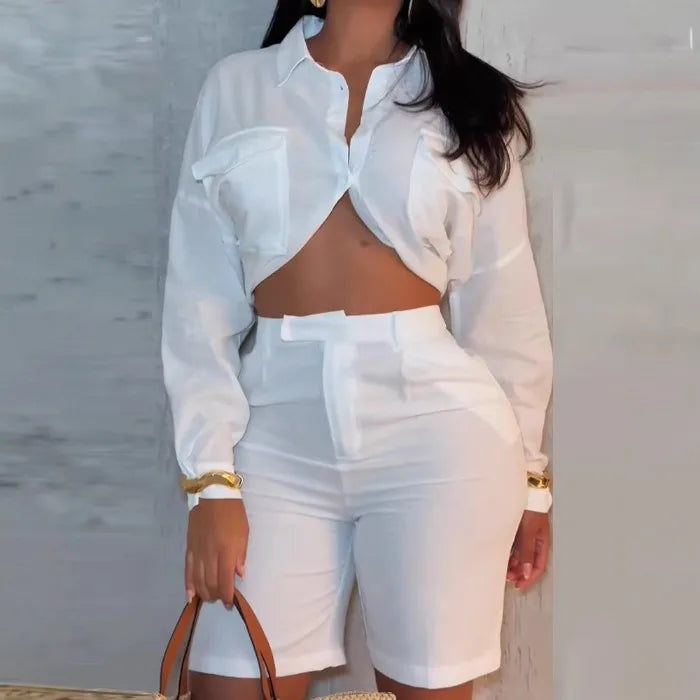 Woman wearing a white chic summer two-piece set with a long-sleeve cropped shirt and high-waisted shorts holding a brown handbag.