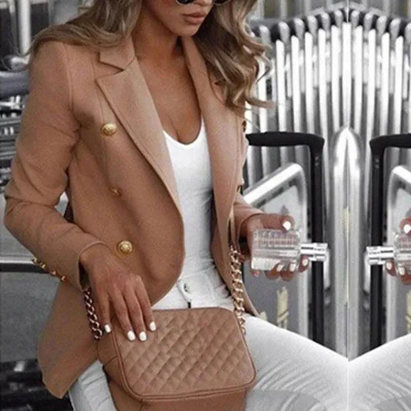 Woman wearing customizable women's blazer suit in camel color with gold buttons, paired with white top and pants, holding quilted handbag and clear glass, seated indoors.