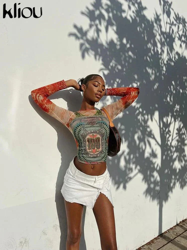 Kliou Dollar Print Long Sleeve Tee with colorful graphic design worn by a woman paired with a white mini skirt against a shadowed wall background