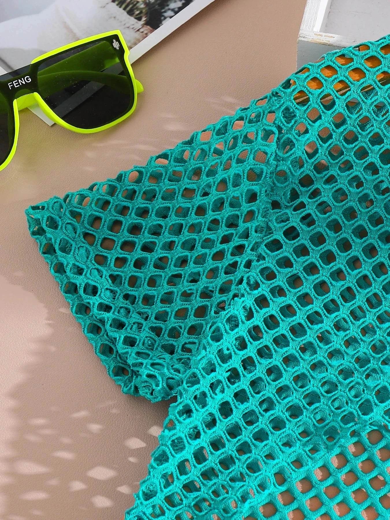 Close-up of turquoise breathable mesh fabric sleeve from girls' trendy 3-piece summer outfit set with neon green sunglasses on beige surface